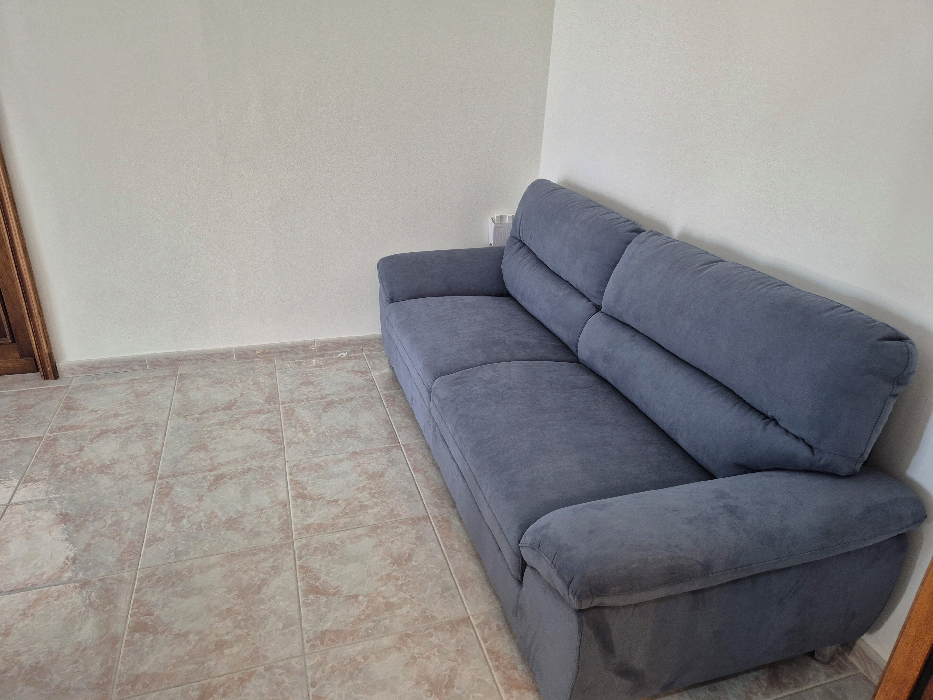 Ein Eckzimmer mit grauem Sofa, gefliestem Boden und einer Holztürleiste.