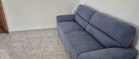 Habitación en esquina con un sofá gris frente a una pared, piso de baldosas y un marco de puerta de madera.