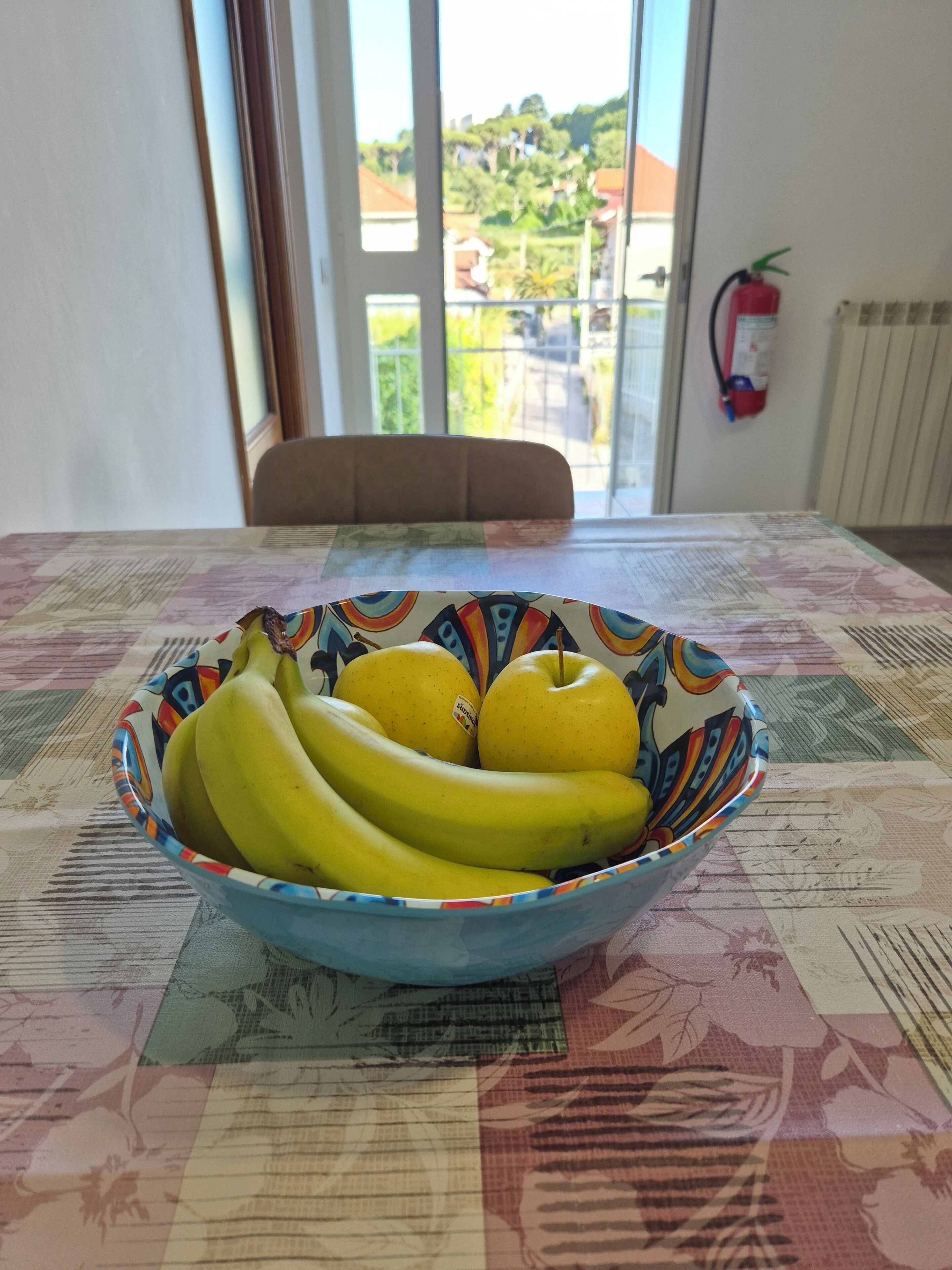 A table with a bowl of bananas on a patterned tablecloth.