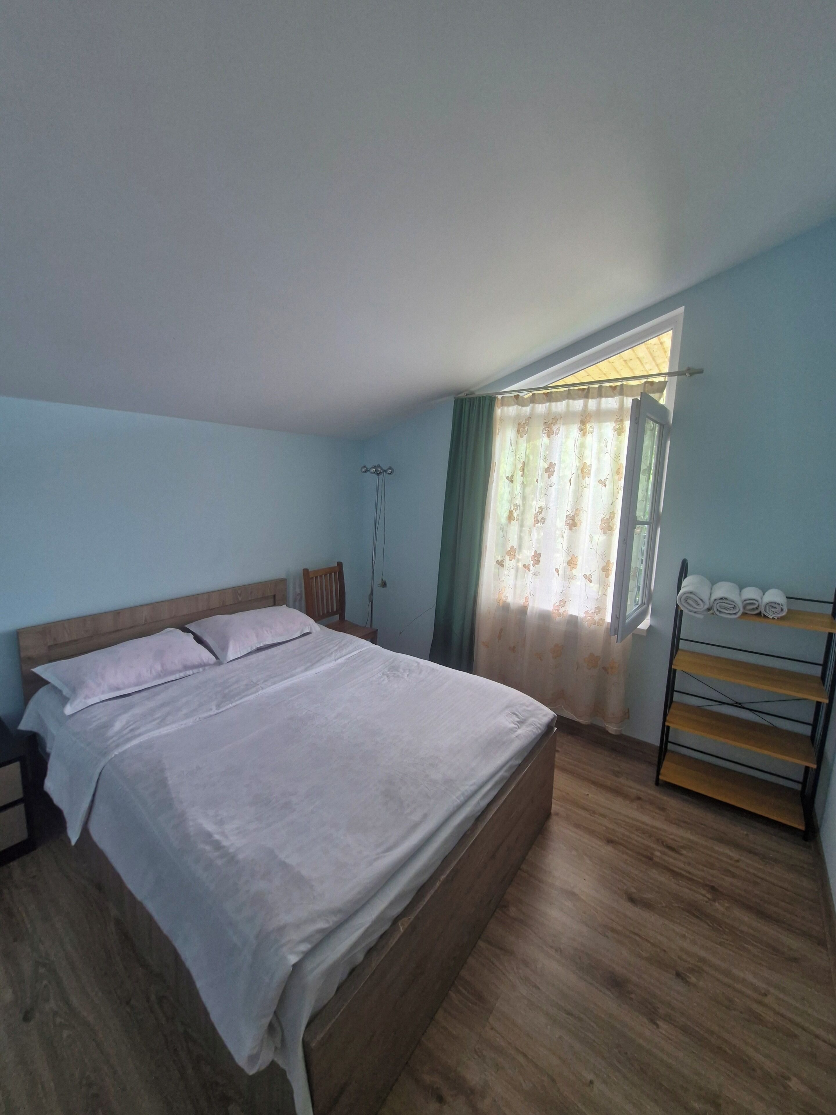 Une chambre à coucher avec un lit en bois, une table de chevet, une fenêtre avec des rideaux et une étagère avec des serviettes.