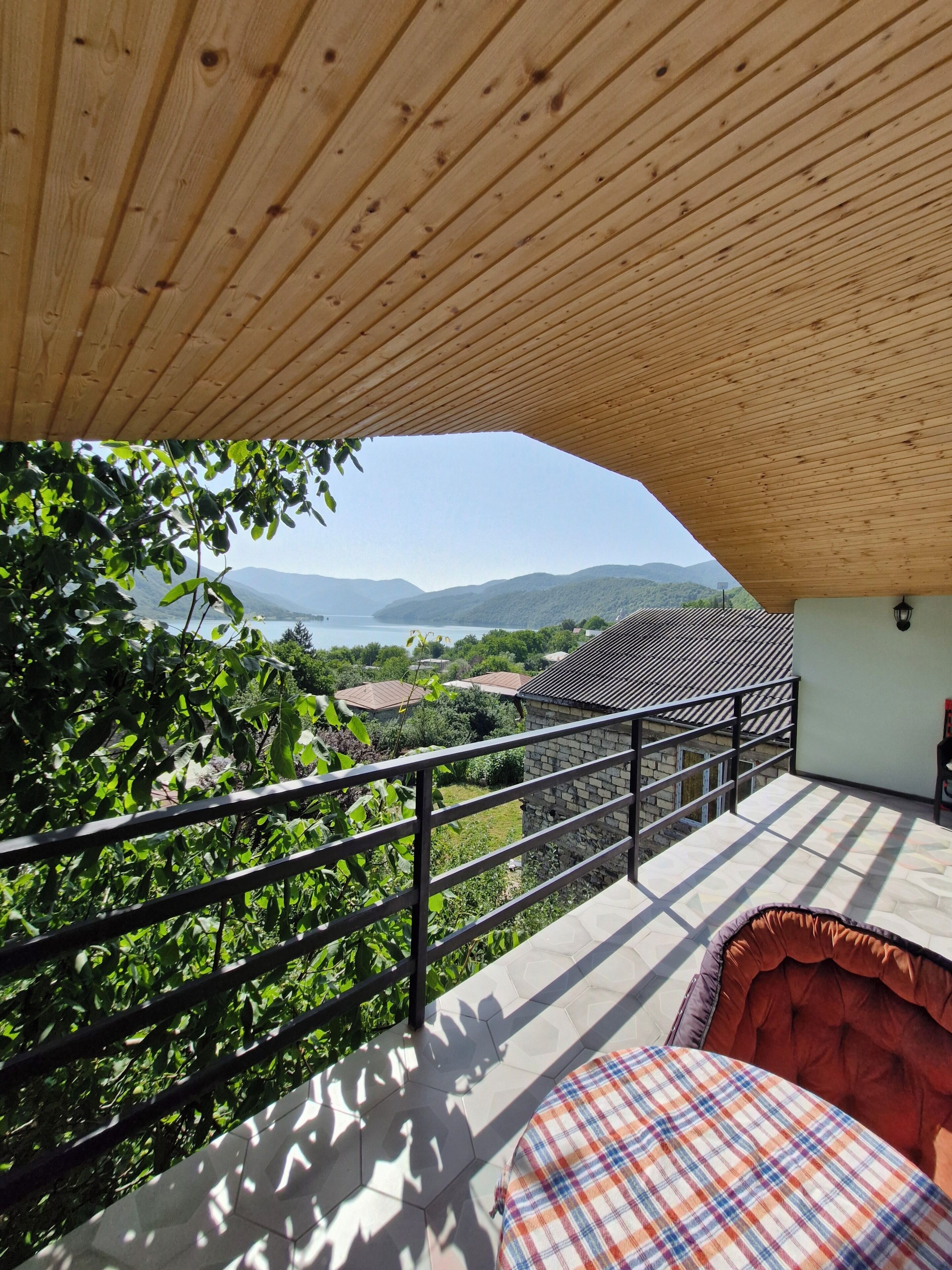 A balcony with a view of a lake and mountains, a wooden ceiling, and a checkered cushion.