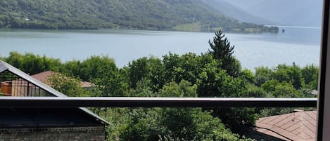 Vista de un lago con montañas al fondo, un balcón con barandilla y algo de vegetación en primer plano.