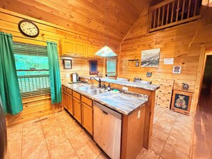 A wooden cabin kitchen with a built-in dishwasher, a sink, and a window with curtains. - 2 King Suites + Loft with 4 Full Beds | Hot Tub | Resort Pool (Sevierville)
