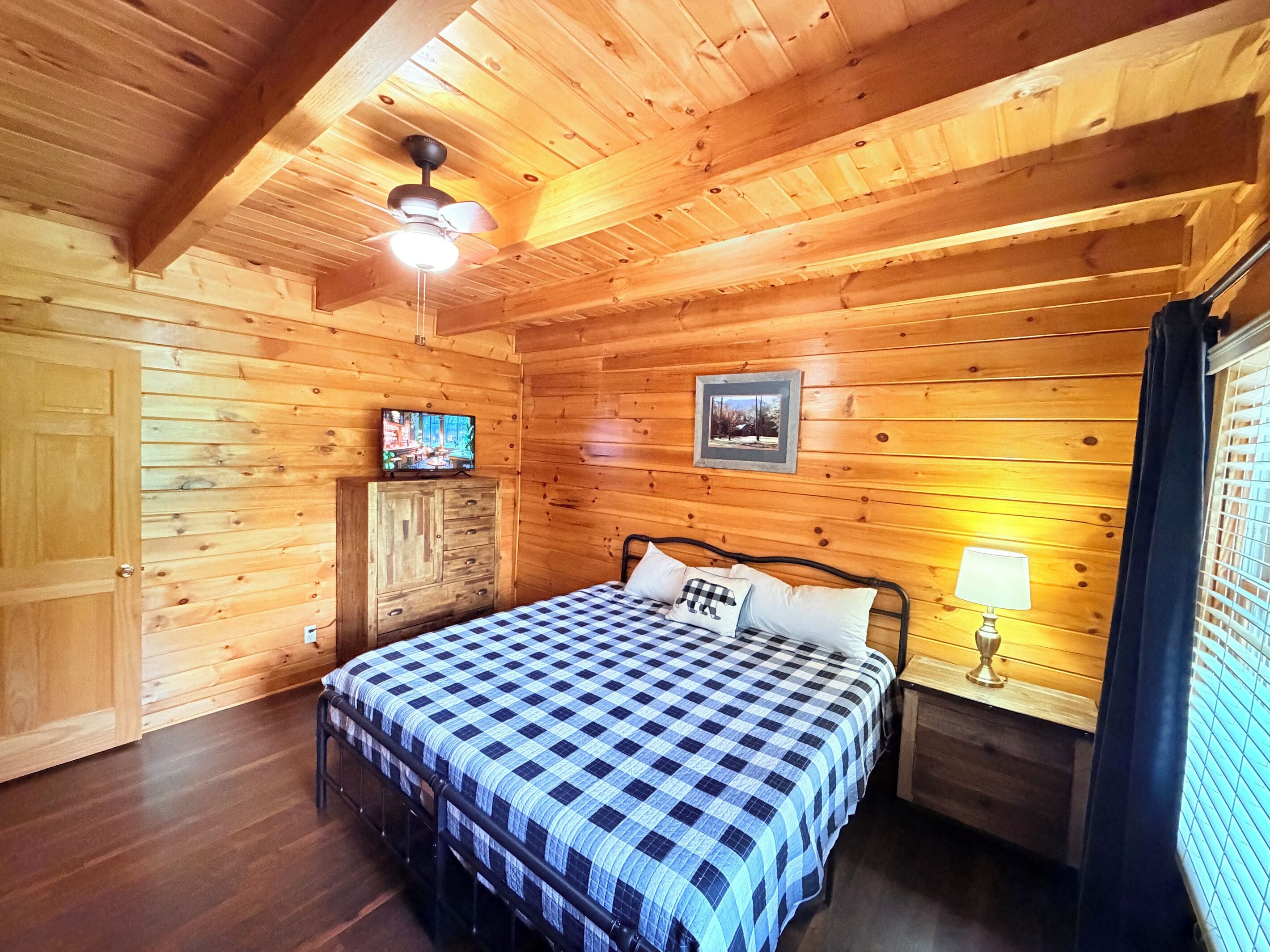 A wooden cabin room with a bed, a television, a framed picture, and a window with curtains.