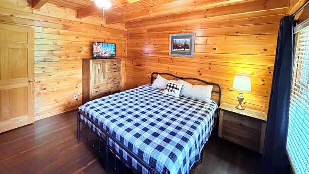 Quarto de cabana de madeira com uma cama, uma televisão, um quadro e uma janela com cortinas.