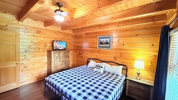 A wooden cabin room with a bed, a television, a framed picture, and a window with curtains.