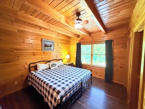 A wooden cabin bedroom with a bed, a ceiling fan, a window with curtains, and a framed picture on the wall. - 2 King Suites + Loft with 4 Full Beds | Hot Tub | Resort Pool (Sevierville)