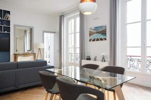 A modern living room with a glass dining table, grey sofa, and large windows.