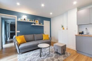 A modern living room with a grey sofa, a round table, and a small ottoman. There is a kitchen area with white cabinets and a countertop. - Renovated apartment between the Opera and the Louvre (Île-de-France)