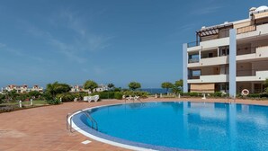A swimming pool with a tiled deck, surrounded by a multi-story building with balconies and a clear view of the sea. - El Rincon 12 - One Bed (Los Cristianos)