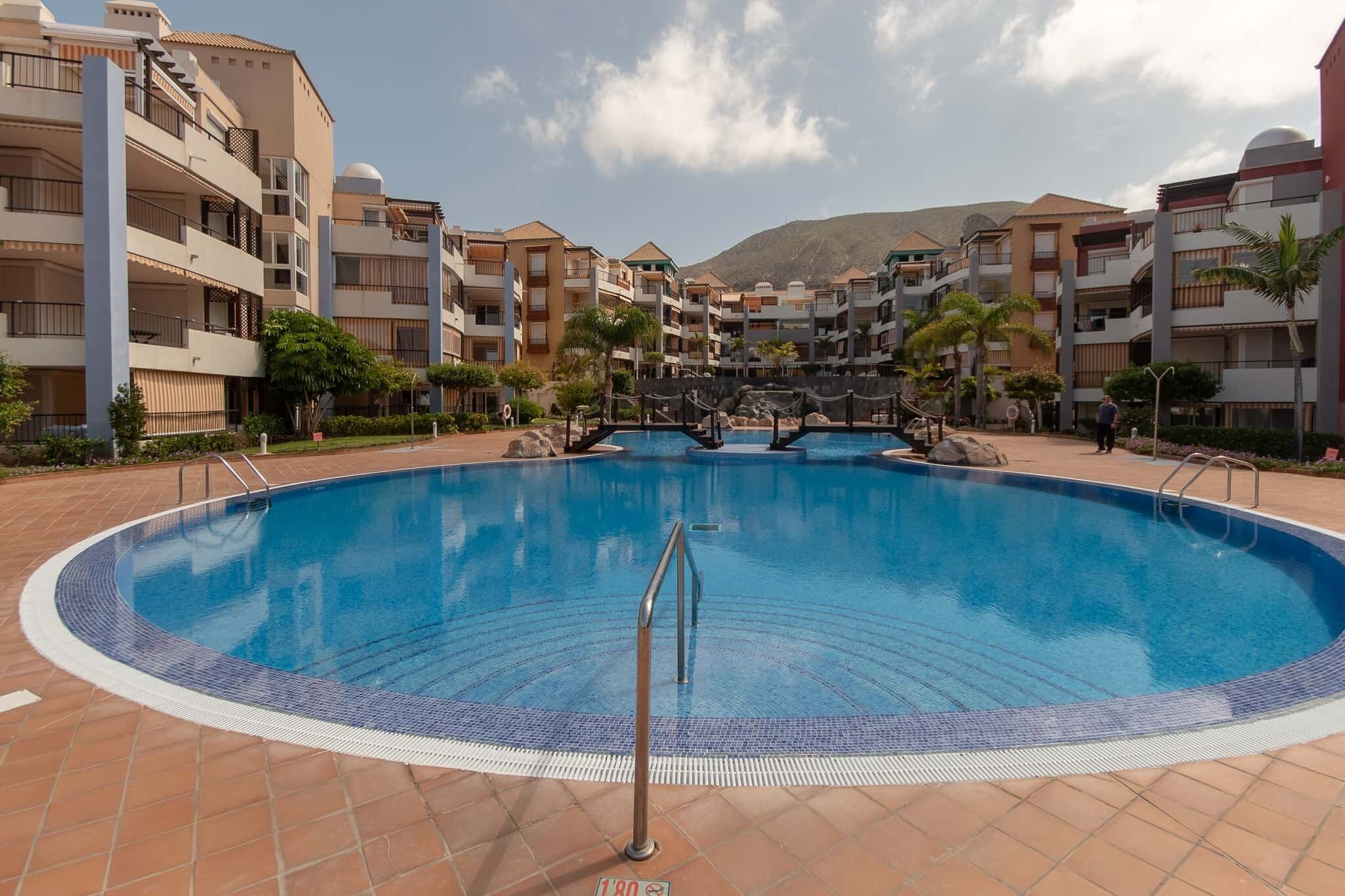 A large circular pool surrounded by multi-story residential buildings with balconies and a mountainous backdrop.
