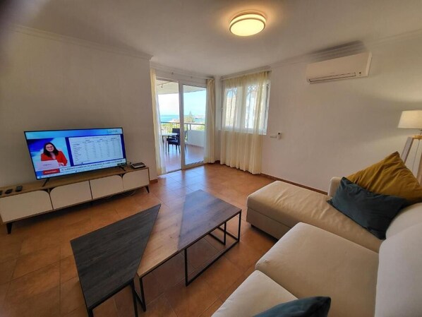 A living room with a television, a sofa, a coffee table, and a sliding glass door leading to a balcony. - El Rincon 12 - One Bed (Los Cristianos)