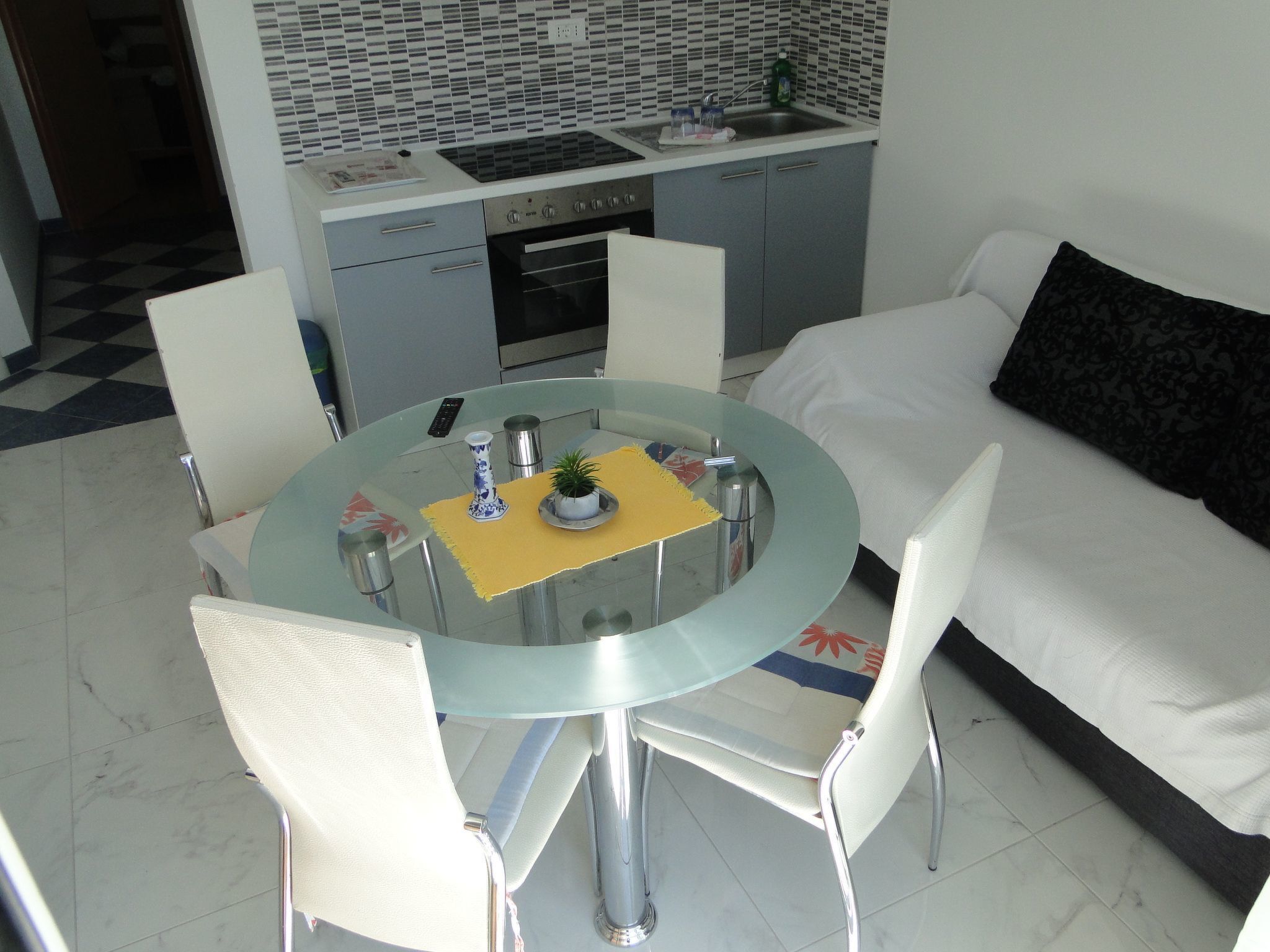 A compact living space with a dining area featuring a round glass table, four chairs, a sofa, and a kitchen with a tiled backsplash.