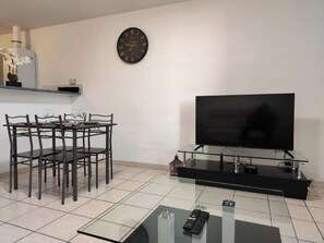 A modern living room with a dining table, chairs, a flat-screen TV on a glass stand, a wall clock, and a small plant. - Panoramic View of the Medieval City - Air Conditioning - 4 People (Carcassonne)