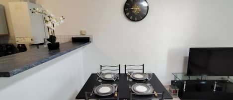 A dining area with a black table set for four, a wall-mounted clock, and a flat-screen TV on a stand.