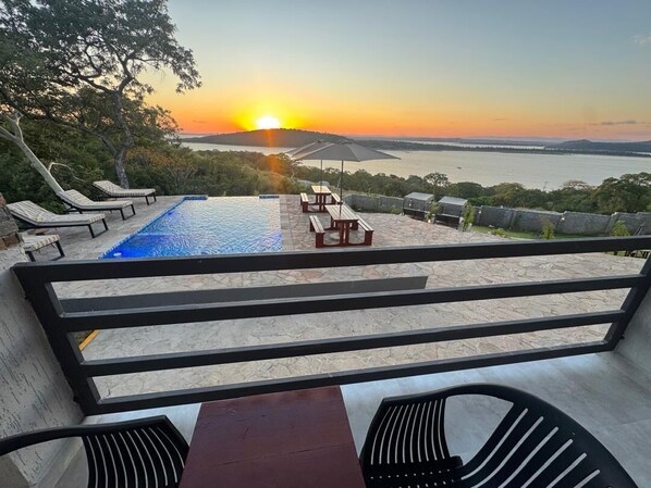 A pool area with lounge chairs and a table, overlooking a body of water and a sunset.
