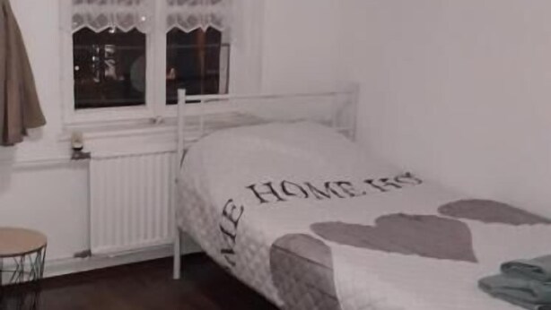 Quarto com cama, janela com cortinas, radiador e tapete xadrez.