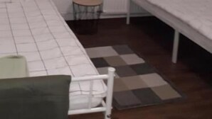 Quarto com cama, uma mesinha e janela com cortinas de renda.