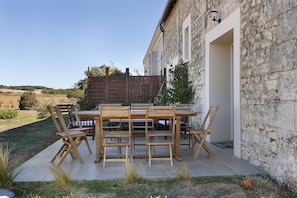 An outdoor patio with a wooden table and chairs, a stone wall, and a view of the countryside.