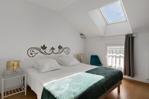 Une chambre avec un puits de lumière, un lit avec une literie blanche, un plaid vert, une table de chevet avec une lampe et une chaise.