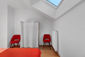 A room with a skylight, two red chairs, a white curtain, and a bed with a red cover.