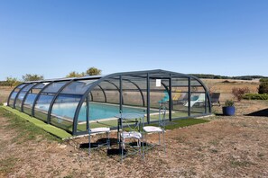 A transparent greenhouse structure with a pool inside, surrounded by a grassy area with a few chairs and a potted plant.