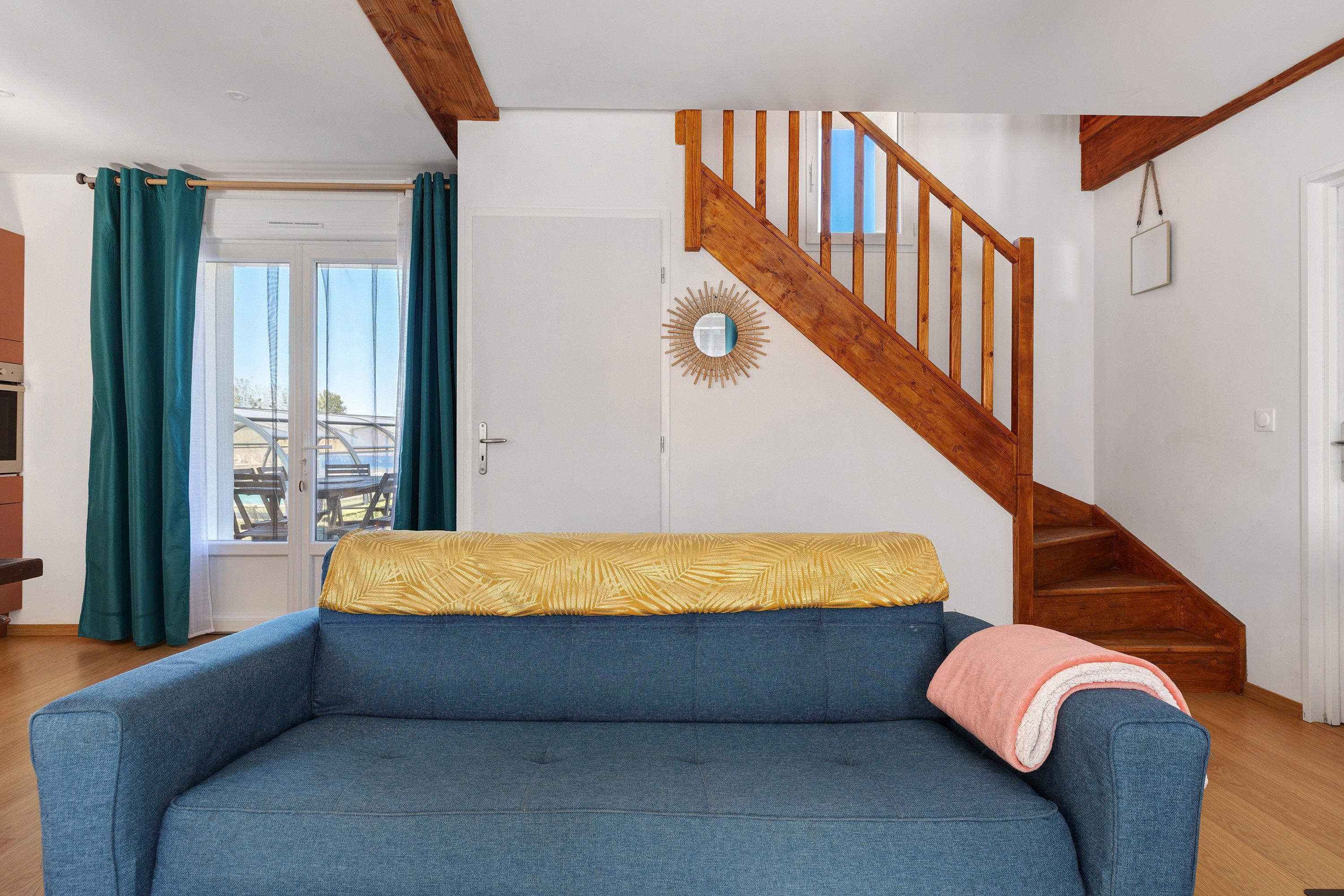 A living room with a blue sofa, a wooden staircase, and a view through a glass door.