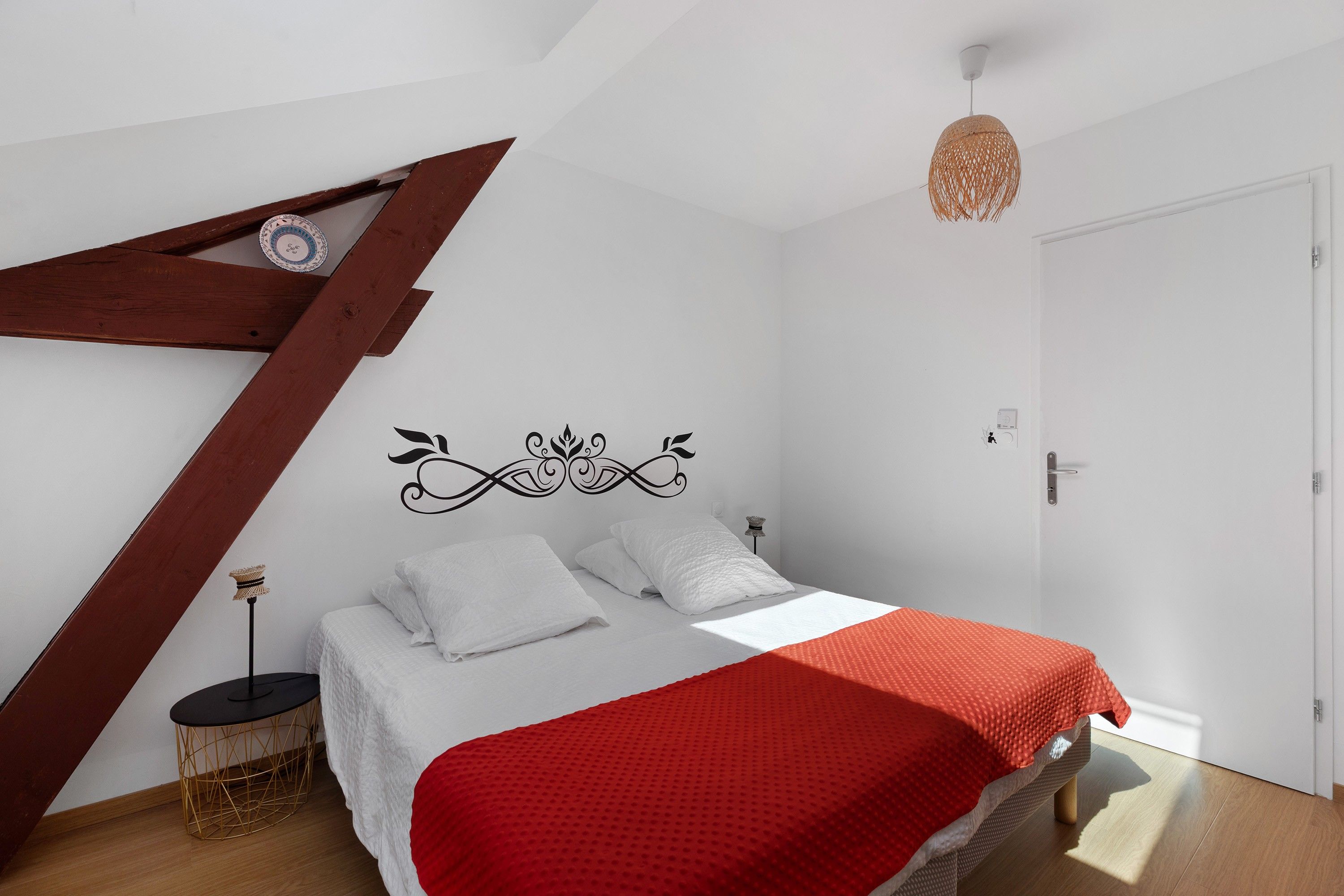 Een slaapkamer met een schuine kap, een bed met witte lakens en een rode deken, een klein rond tafeltje en een decoratief object aan de muur.