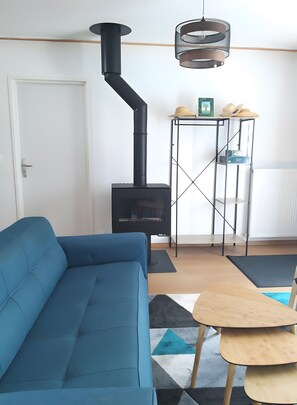 Una sala de estar con un sofá azul, un estufa de leña negra, un estante metálico y una lámpara colgante.