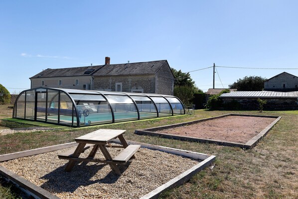 A greenhouse structure with a picnic table in the foreground, set against a backdrop of stone buildings and a clear sky.