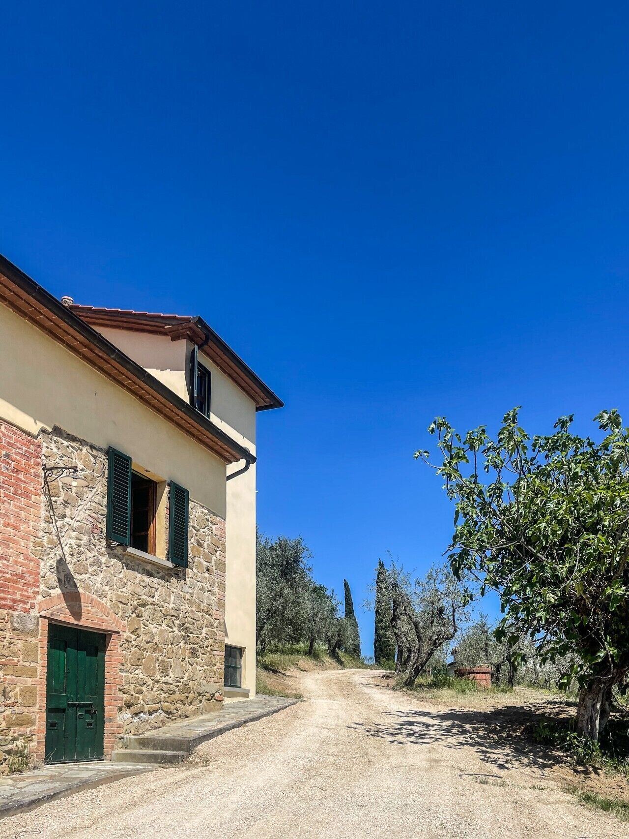 Un chemin de campagne mène à un bâtiment en pierre doté d’une porte verte et de fenêtres, entouré d’arbres et surplombé par un ciel bleu clair.