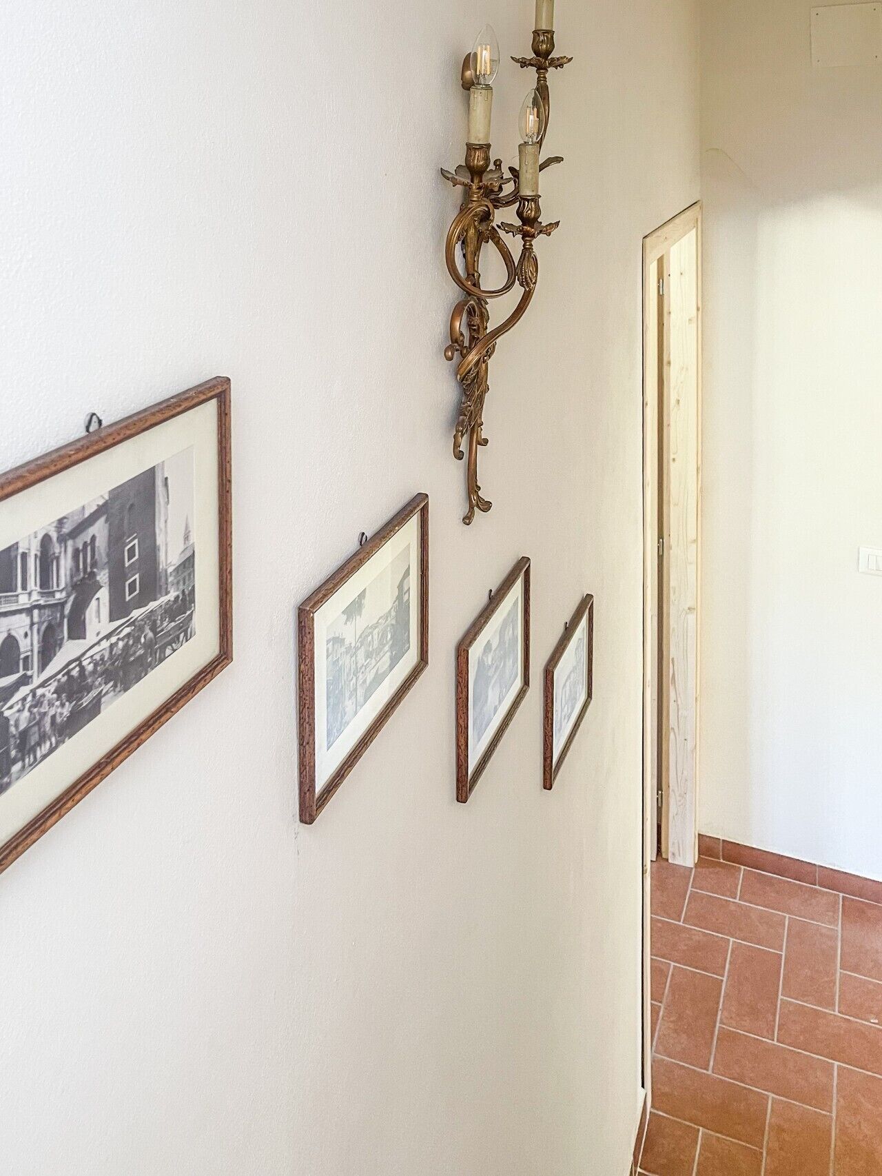 A hallway with framed photographs on the wall, a decorative wall sconce, and a door leading to another room.