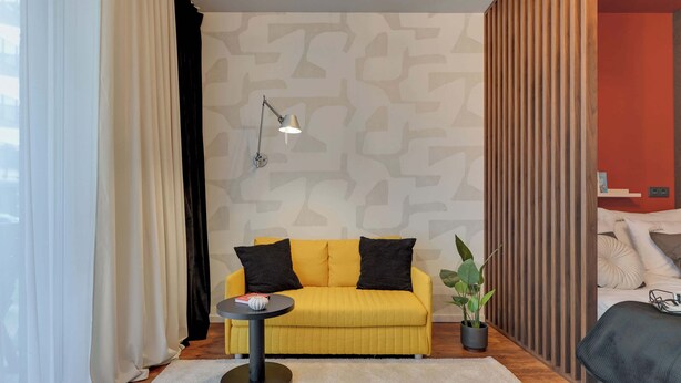En gul sofa med sorte puder, et lille rundt bord med en lampe, en potteplante og en seng med et mørkt tæppe.
