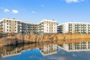 Ein modernes Mehrfamilienhaus mit mehreren Balkonen, einem sich spiegelnden Wasserspiegel davor und blauem Himmel.