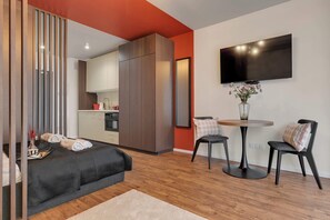 Ein modernes Schlafzimmer mit einem Bett, einem kleinen Tisch mit Stühlen, einem Fernseher und einer Küchenzeile im Hintergrund.