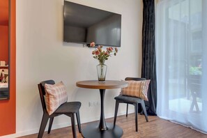 Ein modernes Zimmer mit einem runden Tisch, zwei Stühlen, einem an der Wand befestigten Fernseher und einem Fenster mit transparenten Vorhängen.