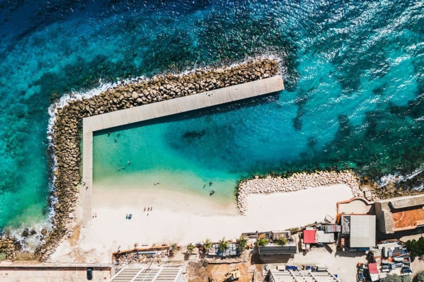 An aerial view of a beach with a clear demarcation of the shoreline, a sandy beach area, and a rocky breakwater.
