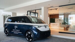 Um carro compacto exibindo a marca 'BOCOBAY LUXURY VACATION RENTAL MANAGEMENT' estacionado em frente a um edifício com fachada de vidro e uma placa que diz 'MAJESTIC APARTMENTS AND HOTEL'.
