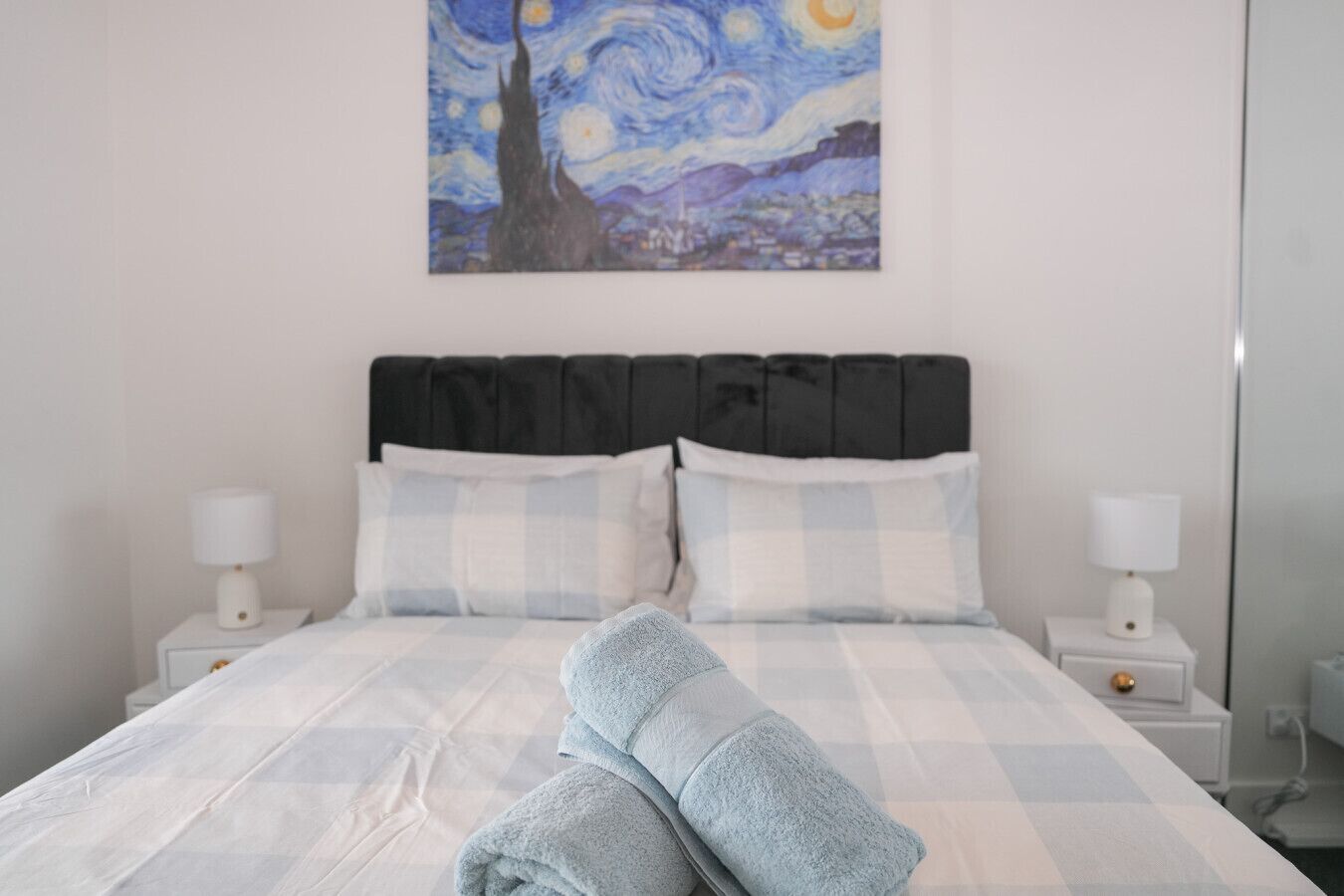 A bed with a checkered duvet, two pillows, and a painting of a night sky above.