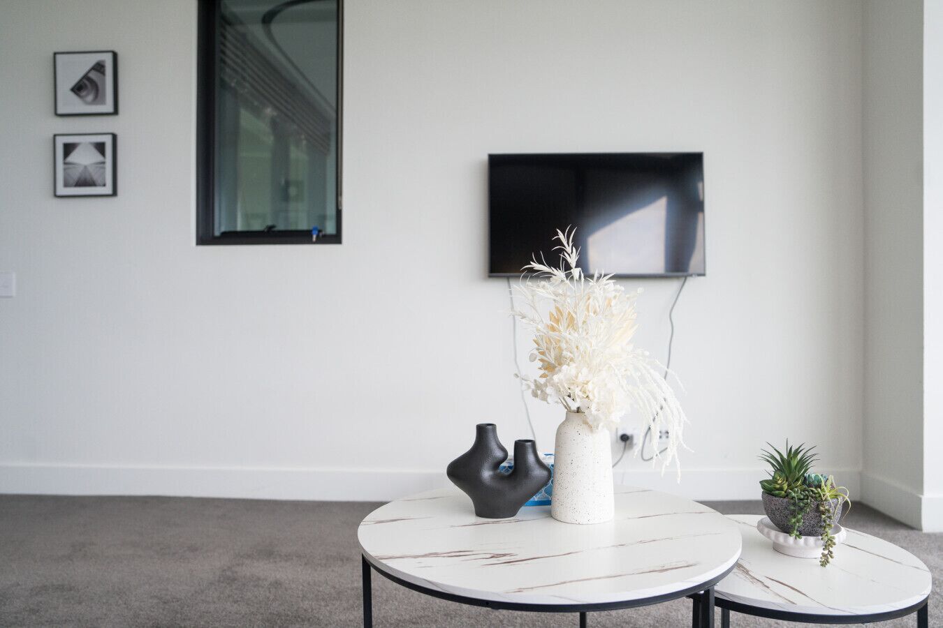 A modern living room with a white wall, a flat-screen TV mounted above a window, and two round tables with decorative vases.