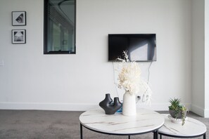 A modern living room with a white wall, a flat-screen TV mounted above a window, and two round tables with decorative vases. - Skyline Retreat in Olympic Park | Views, Parking (Sydney Olympic Park)