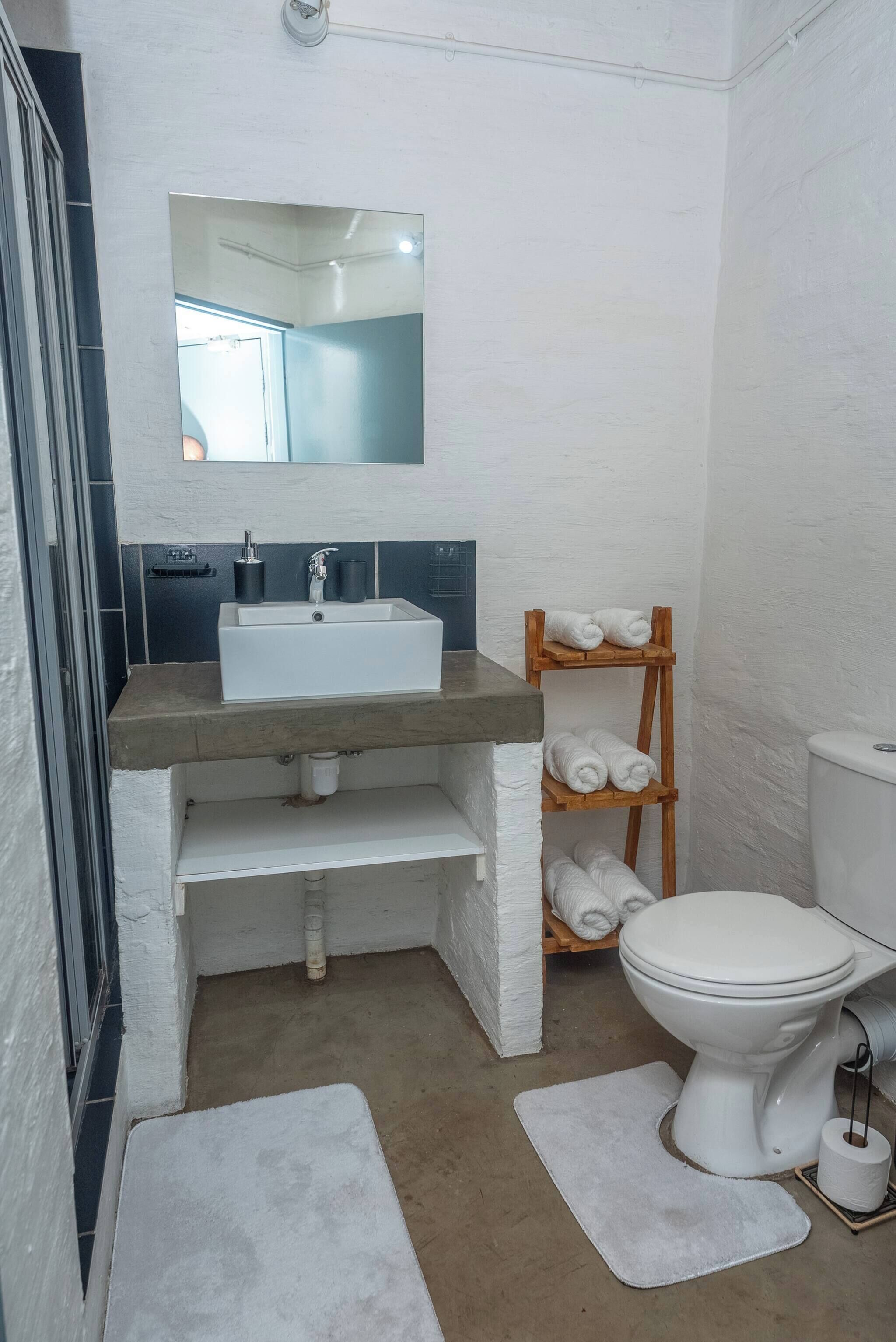 A compact bathroom with a white sink, a mirror above it, a toilet, and a shelf with towels.