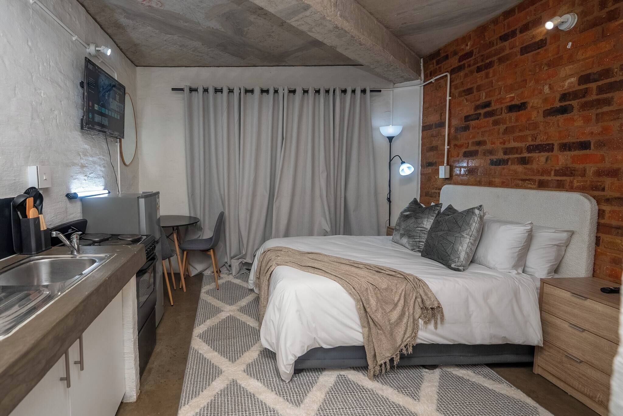 Habitación de hotel pequeña con cama, mesita de noche y una kitchenette.