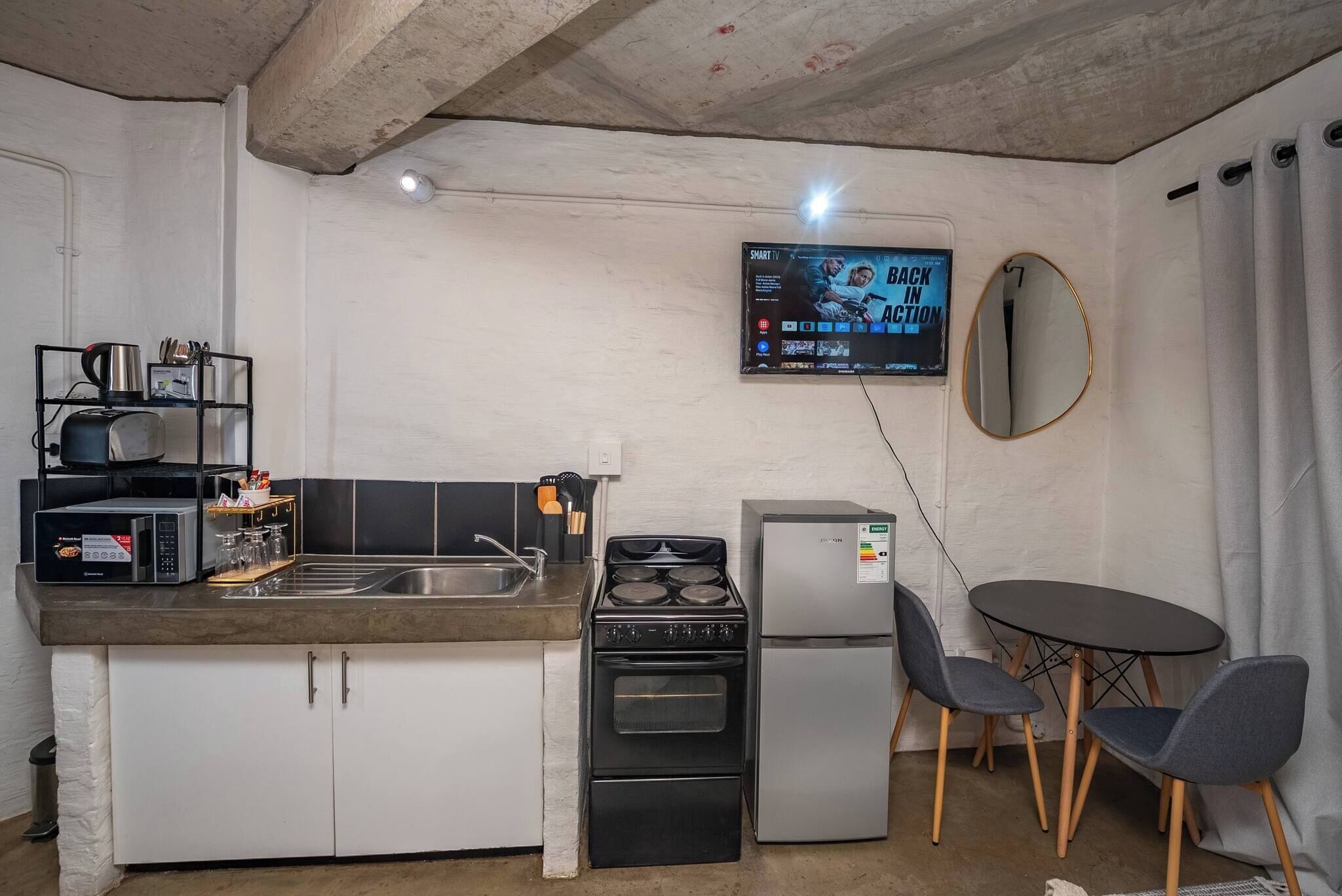 A compact kitchenette with a microwave, toaster, sink, stove, and refrigerator. A flat-screen TV is mounted on the wall, and there is a round mirror above the sink.
