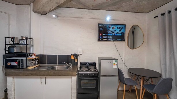 A compact kitchenette with a microwave, toaster, sink, stove, and refrigerator. A flat-screen TV is mounted on the wall, and there is a round mirror above the sink. - The 260 Collection Luxury City Apartment (Johannesburg)