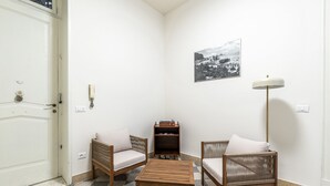Ein Eckzimmer mit Schachbrettfußboden, zwei Korbstühle mit Kissen, ein kleiner Holztisch, ein Bücherregal, eine Wandlampe und eine Tür mit Griff.