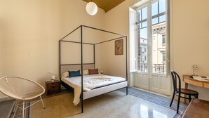 Ein Schlafzimmer mit einem Himmelbett, einem Schreibtisch mit Stuhl, einem Fenster mit Blick auf Gebäude und einer hängenden Leuchte.