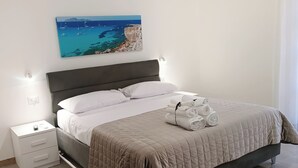 A neatly made bed with a quilted cover, a bedside table with a lamp, and a wall-mounted painting of a coastal scene. - Villa Rosalia with Heated Private Jacuzzi & Sunset View (Tonnara di Bonagia VALDERICE)