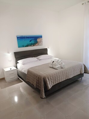 Ein ordentlich bezogenes Bett mit gesteppter Decke, ein Nachttisch mit Lampe und ein Wandbild, das eine Küstenlandschaft zeigt.