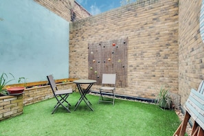 Eine Terrasse mit Kunstgrasanlage, einem Tisch, zwei Stühlen und einer Wand mit hängenden Dekorationen.